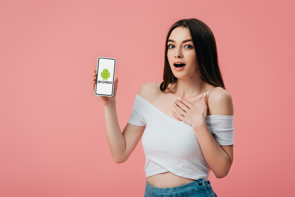 KYIV, UKRAINE - JUNE 6, 2019: beautiful shocked girl holding smartphone with Android logo isolated on pink - Fotografie, Obrázek
