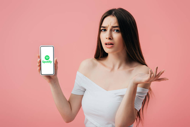 KYIV, UCRAINA - 6 GIUGNO 2019: bella ragazza confusa che tiene lo smartphone con l'app Spotify e mostra un gesto di spalluccio isolato sul rosa
 - Foto, immagini