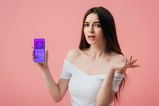 gyönyörű zavaros lány gazdaság okostelefon online vásárlás app és bemutatja vállrándítással gesztus elszigetelt rózsaszín - Fotó, kép