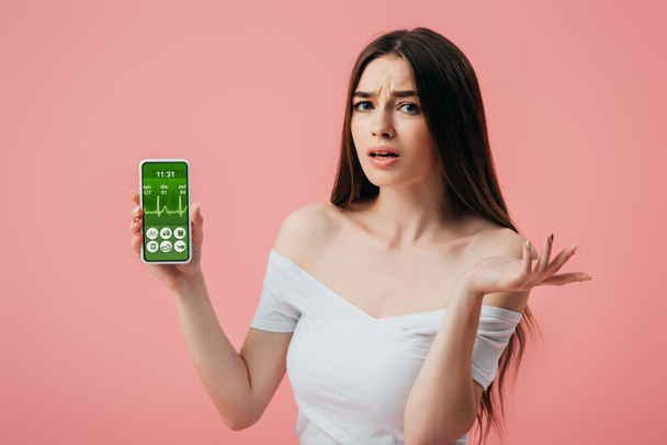 gyönyörű zavaros lány gazdaság okostelefon egészséges életmód app és bemutatja vállrándítással gesztus izolált rózsaszín - Fotó, kép
