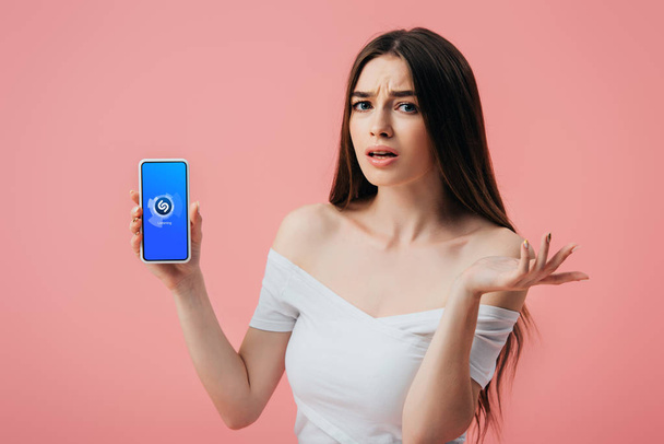 KYIV, UKRAINE - JUNE 6, 2019: beautiful confused girl holding smartphone with Shazam app and showing shrug gesture isolated on pink - Foto, imagen