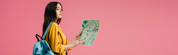 Panoramaaufnahme eines verträumten Mädchens mit Rucksack, das Karte in der Hand hält und isoliert auf rosa - Foto, Bild