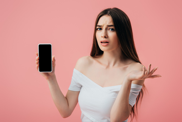 白い画面でスマートフォンを保持し、ピンクに隔離されたシュラッグジェスチャーを示す美しい混乱した女の子 - 写真・画像