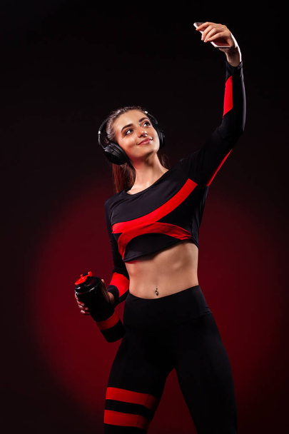 Beautiful young and fit woman in sportswear and headphones listening music and taking a selfie. Isolated on black background. - 写真・画像