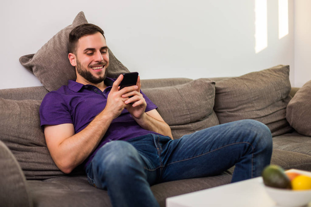 Young man man using mobile phone while sitting on sofa  in his living room. - Foto, Imagen