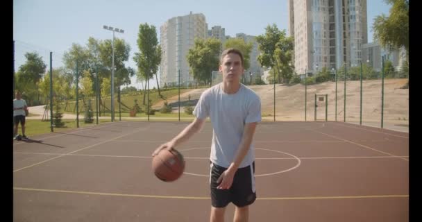 Closeup portrait of young caucasian male basketball player throwing a ball in the hoop on the court with cityscapes on the background - Кадры, видео