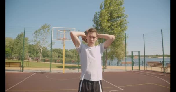 Closeup portrait of young caucasian male basketball player holding a ball behind back looking at camera confidently on the court outdoors with bridge on the background - Video