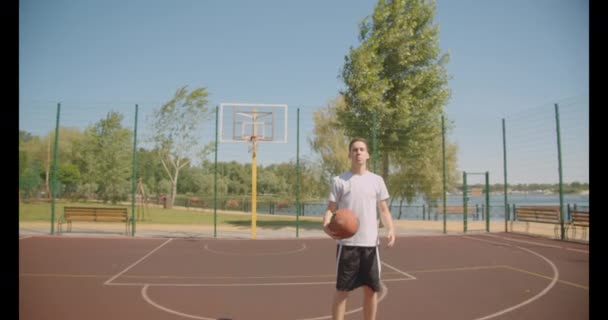 Closeup portrait of young caucasian male basketball player holding a ball behind back looking at camera on the court outdoors with bridge on the background - 映像、動画