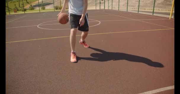 Closeup portrait of young attractive caucasian male basketball player throwing a ball in the hoop on the court with houses on the background - Materiaali, video