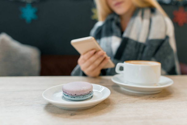 Macaroon in plate, cup of coffee, beautiful blonde girl in woolen blanket with smartphone - 写真・画像