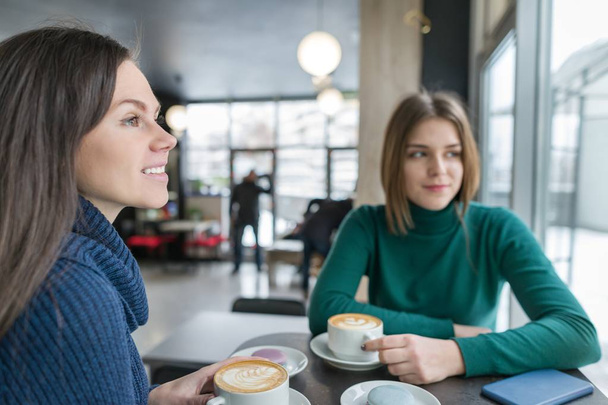 Two smiling young women in coffee shop with coffee art and macaroons in plates, girls talking, winter season wearing warm sweaters - Foto, imagen