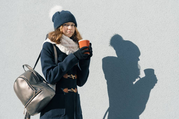 Winter sunny outdoor portrait of teenage girl wearing a coat and knit hat with cup of hot drink, copy space - Fotó, kép