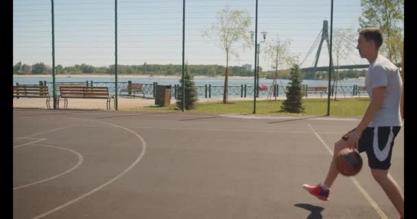 Closeup portrait of young sporty caucasian male basketball player throwing a ball in hoop on the court outdoors with bridge on the background - Materiaali, video