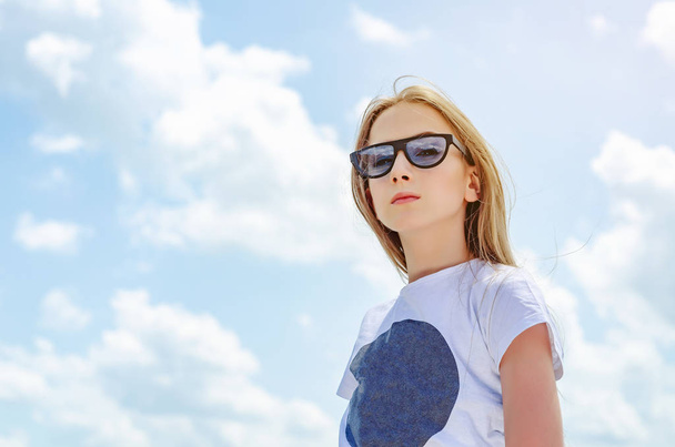 Summer portrait of a blonde girl in sunglasses on a background of the sky with clouds - Photo, Image