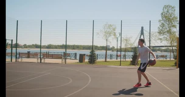 Closeup portrait of young athletic caucasian male basketball player throwing a ball in hoop on the court outdoors with bridge on the background - Záběry, video