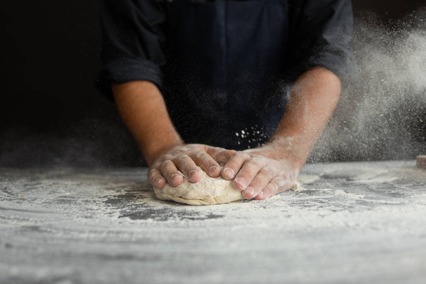 chef kneads raw dough, dust from flour flies in different directions, male hands on a black background - Photo, Image