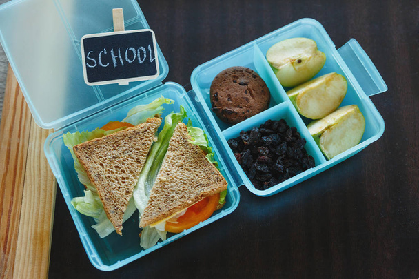 自家製サンドイッチ、緑のリンゴ、クッキー、鉛筆、時計、テーブルの上にノートを持つ学校の青いランチボックス。学校で健康的な食事。学校の背景に戻る。フラットレイ、トップビュー. - 写真・画像