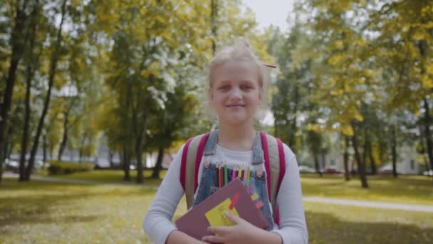 Joyful schoolgirl with backpack holding a book staying in the school yard. Portrait of a pretty smart girl smiling outdoors. Slow Motion Shot. - Video