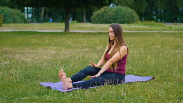Fiatal kaukázusi nő pihen a gyakorló jóga szabadtéri helyen reggel. Gyönyörű nő csinál jóga a parkban a zöld fű - Felvétel, videó