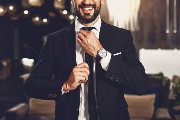 Ένας νεαρός με αυτοπεποίθηση που αγγίζει τη γραβάτα του και χαμογελάει ενώ στέκεται μόνος - Φωτογραφία, εικόνα