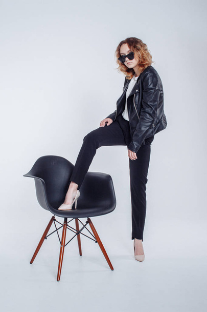 Pretty young fashion sensual woman posing on white wall background with black chair dressed in hipster style leather jacket and sunglasses outfit. Stylish fashionable girl hipster in black clothes. - Fotoğraf, Görsel