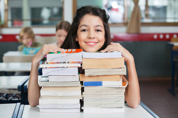 Happy Schoolgirl Resting Chin On Stacked Books At Desk - Foto, afbeelding