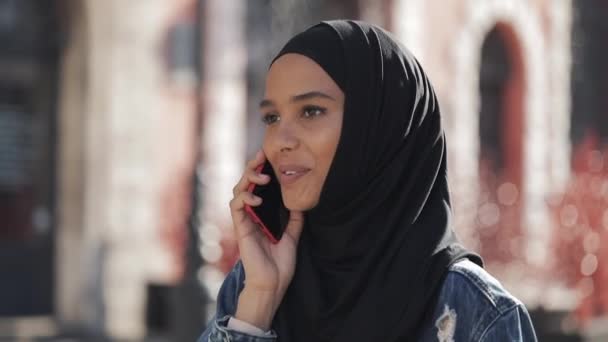 Smiling young muslim woman wearing hijab headscarf talking with friends on a smartphone in the city, a lady walks along old street in black hijab. - Πλάνα, βίντεο