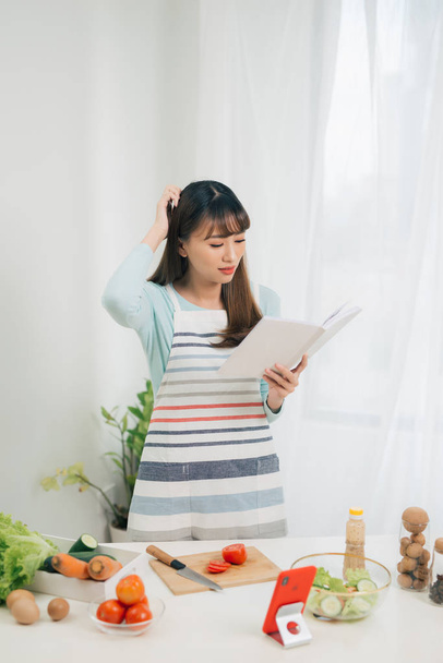 Mujer joven leyendo libro de cocina en la cocina, buscando receta - Foto, imagen
