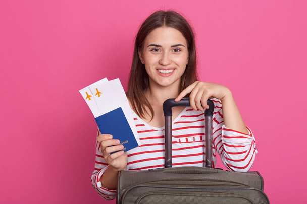 Close up portrait of happy European girl with suitcase, airplane tickets and passport isolated on rosy background, lady feels excited about trip, wearing casual white shirt with red stripes. Traveling - Photo, image