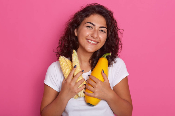 Picture of cheerful attractive model looking directly at camera, holding corn cobs and squash, keeping close to body, having black curly hair, posing isolated over pink background in studio. - Foto, immagini