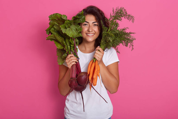 Close up portrait of attractive caucasian smiling woman isolated on rose studio background, wearing white casual t shirt, holding beets and carrots in hands. Raw dood eatinf and healthy eating concept - Zdjęcie, obraz
