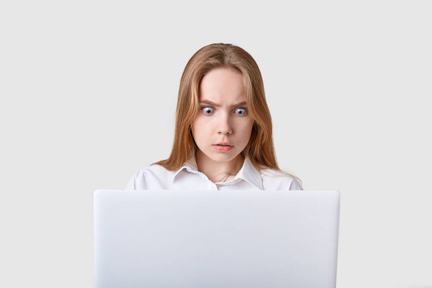 Portrait of shocked woman sitting in front of white laptop with wide opened eyes. Attractive girl looks at computer screen with astonished facial expression, sees somothing terrible, working online. - 写真・画像