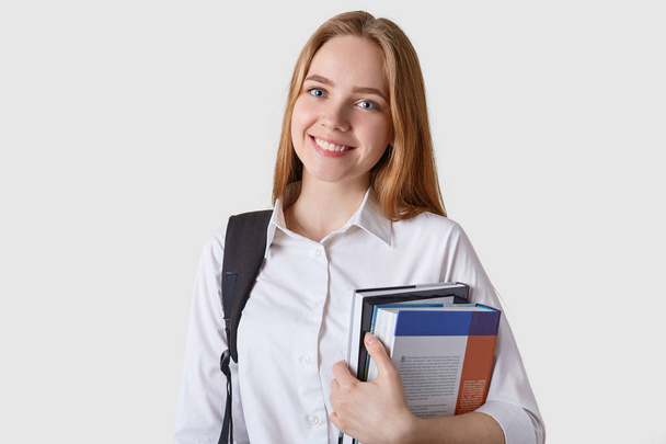 Beltéri shot gyönyörű tinédzser lány, fekete hátizsák és papír mappa a kezében, látszó boldog, vonzó tanuló nő izolált fehér background. Tinédzserek, az oktatás és a siker koncepciója. - Fotó, kép