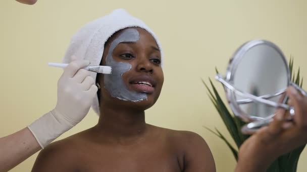 Shooting of cosmetologist hand on applying mask on face of young African-American woman in spa salon - Séquence, vidéo