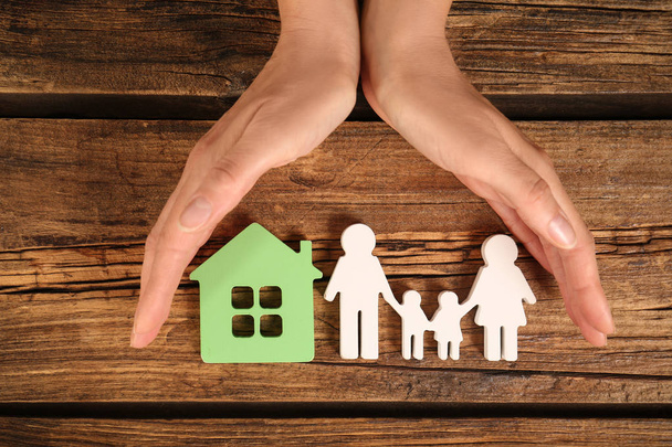 Woman holding hands near figures of house and family on wooden background, top view - Foto, Imagem