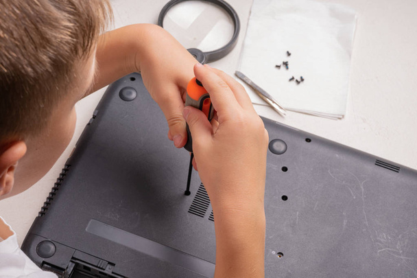 Um menino de 10 anos está classificando um laptop para limpeza e manutenção. Foco seletivo. Chaves de fenda, cilindro de purga, lupa e limpador de pulverização no quadro
. - Foto, Imagem