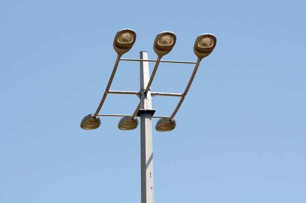 hoher starker Metallmast mit sechs großen modernen Lichtreflektoren, die oben auf klarem blauem Himmelshintergrund montiert sind - Foto, Bild