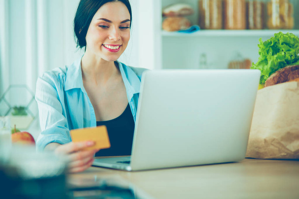 Smiling woman online shopping using computer and credit card in kitchen - Photo, Image