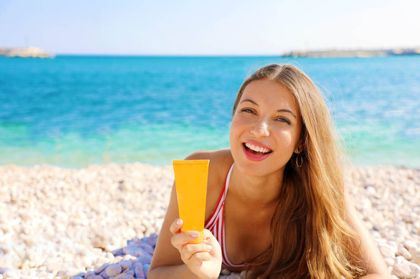 Femme souriante tenant protection tube crème solaire couché sur la plage de galets. Écran solaire fille montrant crème solaire dans un récipient en plastique
. - Photo, image