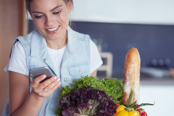 Smiling woman with mobile phone holding shopping bag in kitchen - Photo, Image