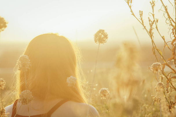 head backof red-haired girl sitting on the ground in a field among dried flowers and enjoys nature at sunset, young woman relaxing, concept of rest, healthcare, harmony, lifestyle - 写真・画像