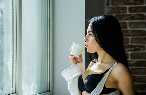 自宅でリラックス魅力的な女性のランジェリー。完璧なコーヒータイム。でも最初のコーヒー静けさと平和クリーミーなカプチーノを一口。朝の儀式窓の近くでコーヒーを飲む女の子のセクシーな胸 - 写真・画像