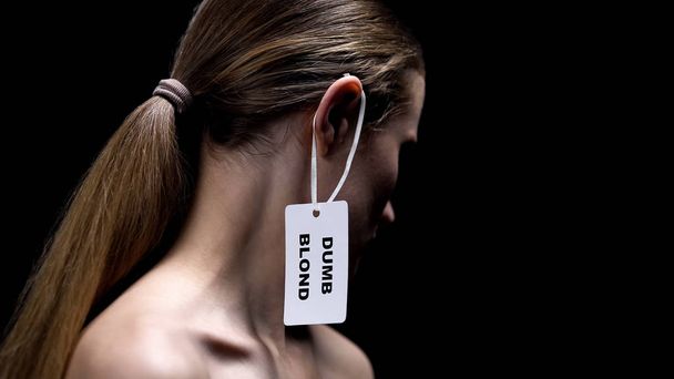 Lady with dumb blonde tag on ear against dark background, humiliation stereotype - 写真・画像