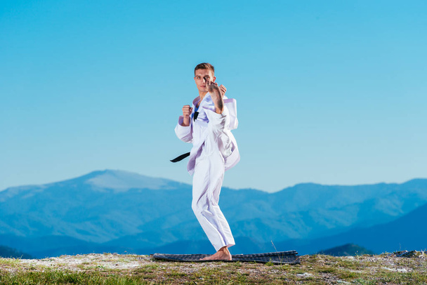 Atleta de karatê loiro faz kata no topo de uma montanha enquanto executa uma linha de chutes, socos e blocos no topo de uma montanha em um dia ensolarado. - Foto, Imagem