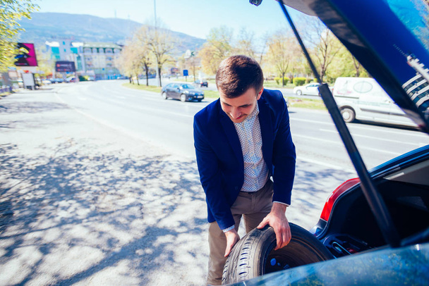 Уставший бизнесмен в солнечный день вытаскивает колесо из машины, чтобы поменять колесо.
. - Фото, изображение