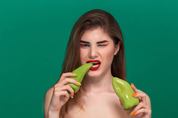 Крупный план портрет красивой молодой женщины с ярким цветом макияжа держа зеленый перец стоя изолированы перед зеленым фоном
 - Фото, изображение