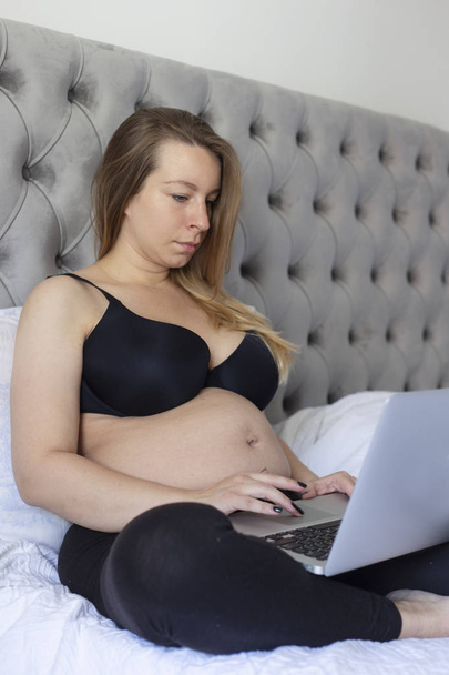 Viehättävä raskaana oleva nainen käyttää kannettavaa tietokonetta istuessaan sängyssä
 - Valokuva, kuva