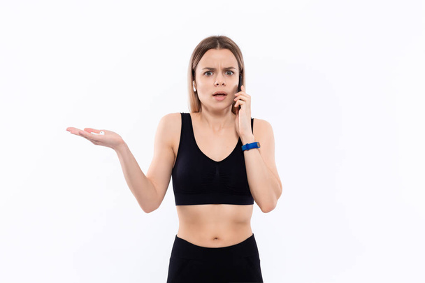 Mujer rubia deportiva joven asombrada en una ropa deportiva negra con auriculares inalámbricos hablando por teléfono de pie sobre fondo blanco
. - Foto, Imagen
