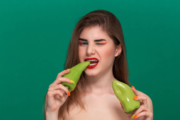 Close-up portret van mooie jonge vrouw met felle kleur make-up houden groene peper staande geïsoleerd Infront van een groene achtergrond - Foto, afbeelding
