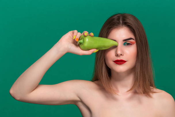 Closeup portrait of beautiful young woman with bright color make-up holding green pepper standing isolated infront of a green background - Photo, image